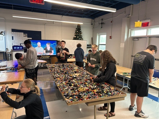 Indoors, individuals gather around a table covered with Legos, engaged in building activities. A television displays content in the background.