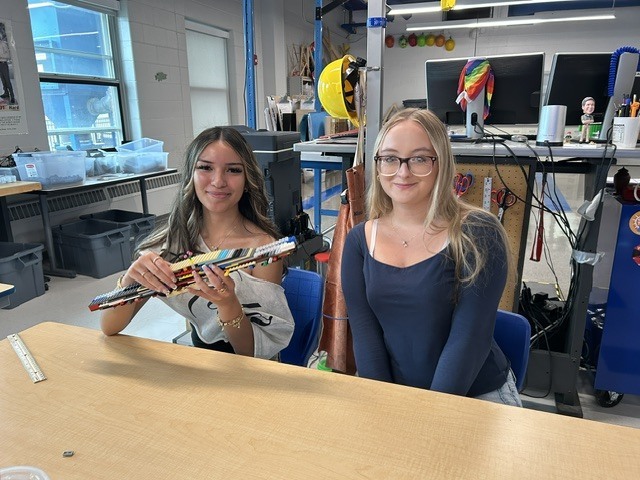 Two girls sit at a desk. One holds a colorful object. Behind them, several storage boxes and a window.