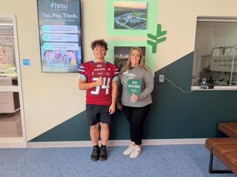 Two people stand near a wall with green and white design. A boy wears a red jersey with the number 34. A woman holds a green sign.