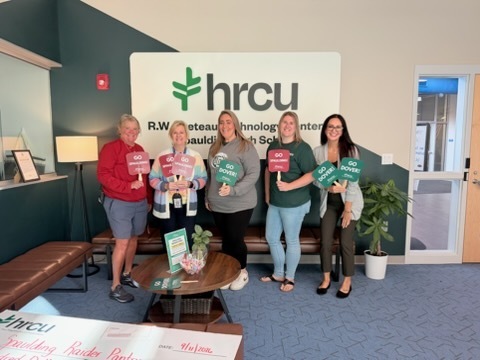 Five women stand together in a room, holding signs, near a table, a couch, and a potted plant.