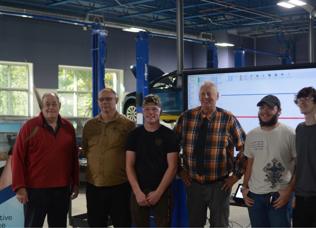 Six men stand in a vehicle repair shop, posing for a photo in front of a car on a lift.