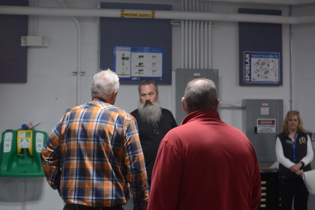 A group of people stands in a room with white walls and gray cabinets. Two individuals converse, while a third person listens.