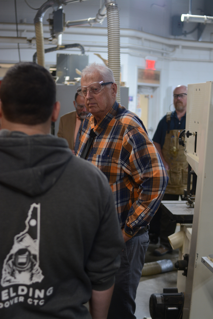A man in a plaid shirt converses with others in a workshop. Behind him, a large machine and a door.