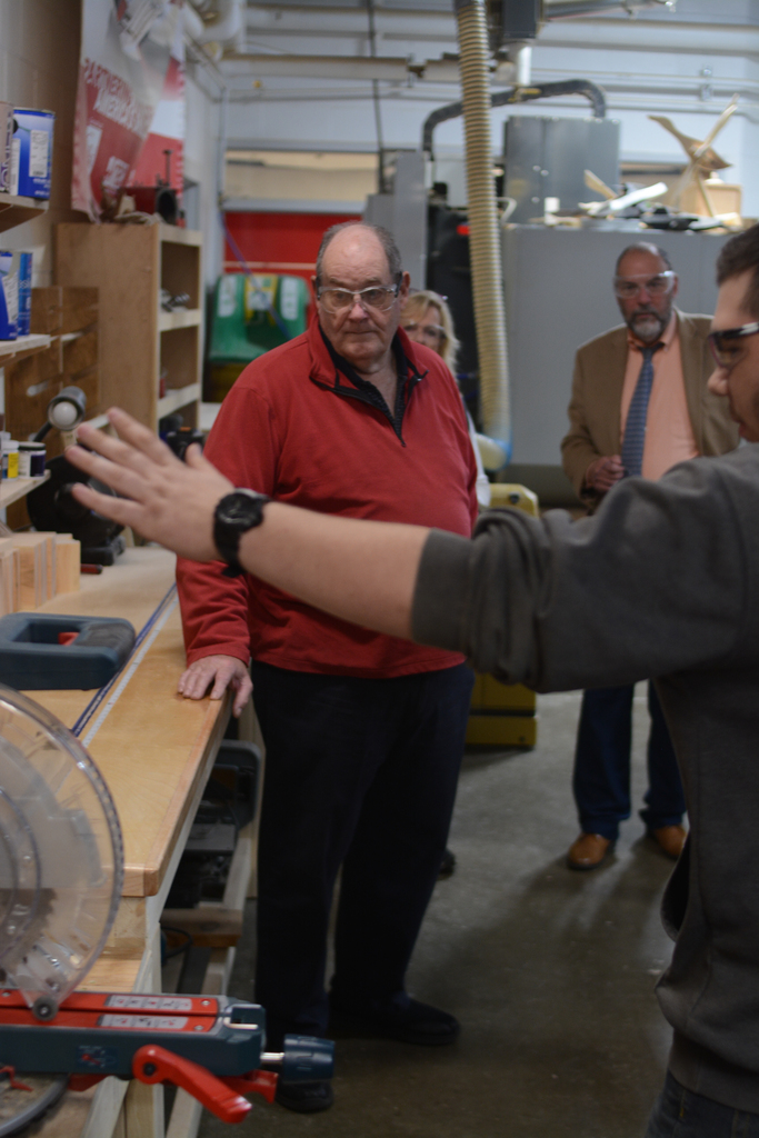 A man explains something to another man in a workshop with several people standing behind them.