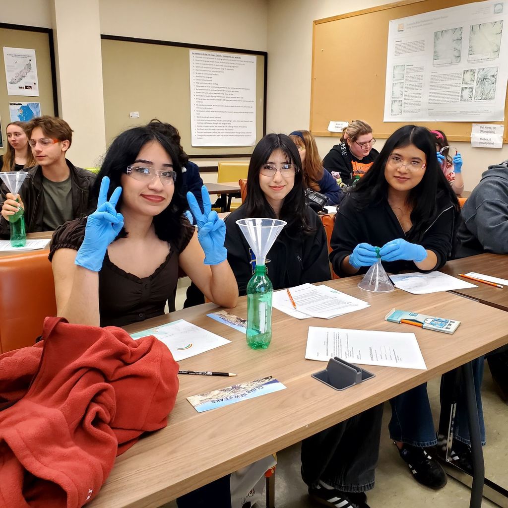 A classroom with several students. Three students are seated in front of a table, smiling, and wearing blue gloves.