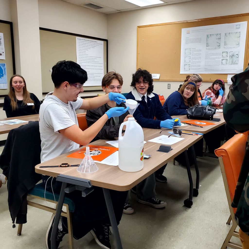 Several students wearing lab coats and gloves work at tables, one pouring liquid from a jug.