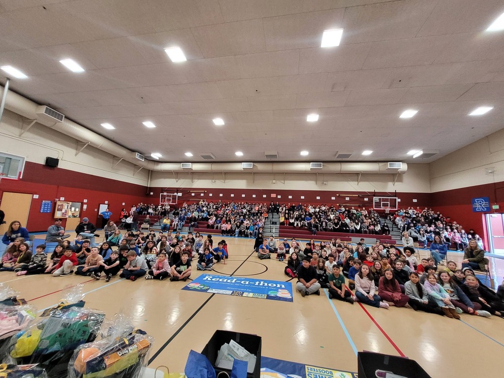A gym with spectators seated in bleachers and on the floor. The floor has bags and boxes.
