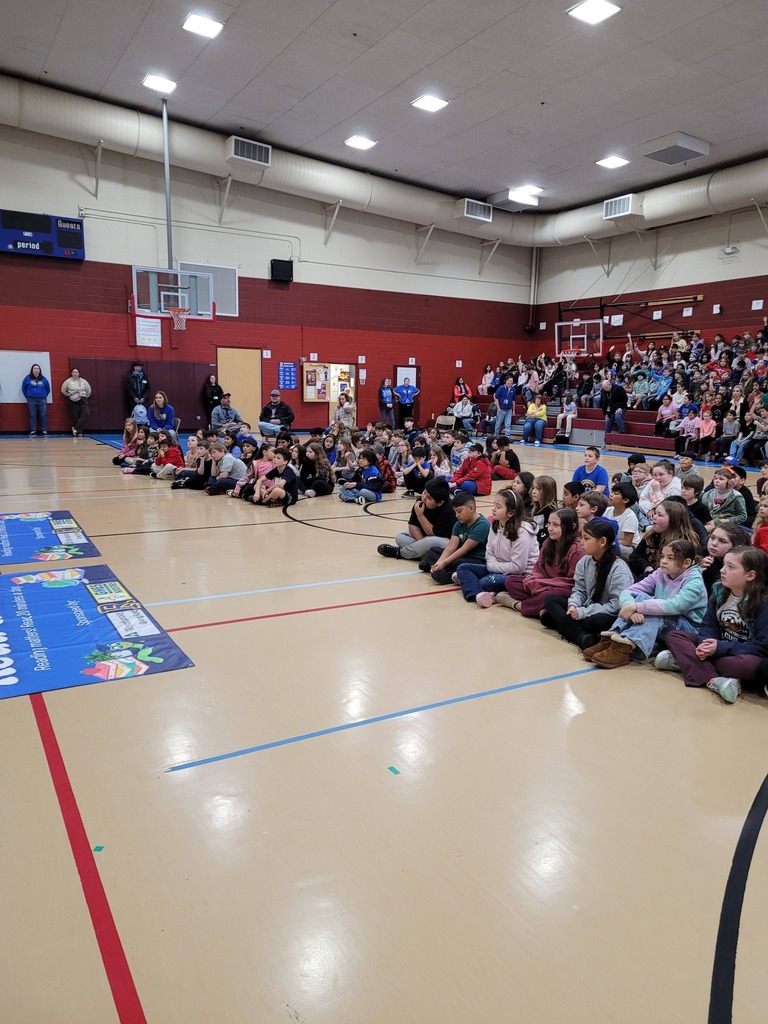 Many people seated on a basketball court, possibly attending an event. Blue banners and a basketball hoop in background.