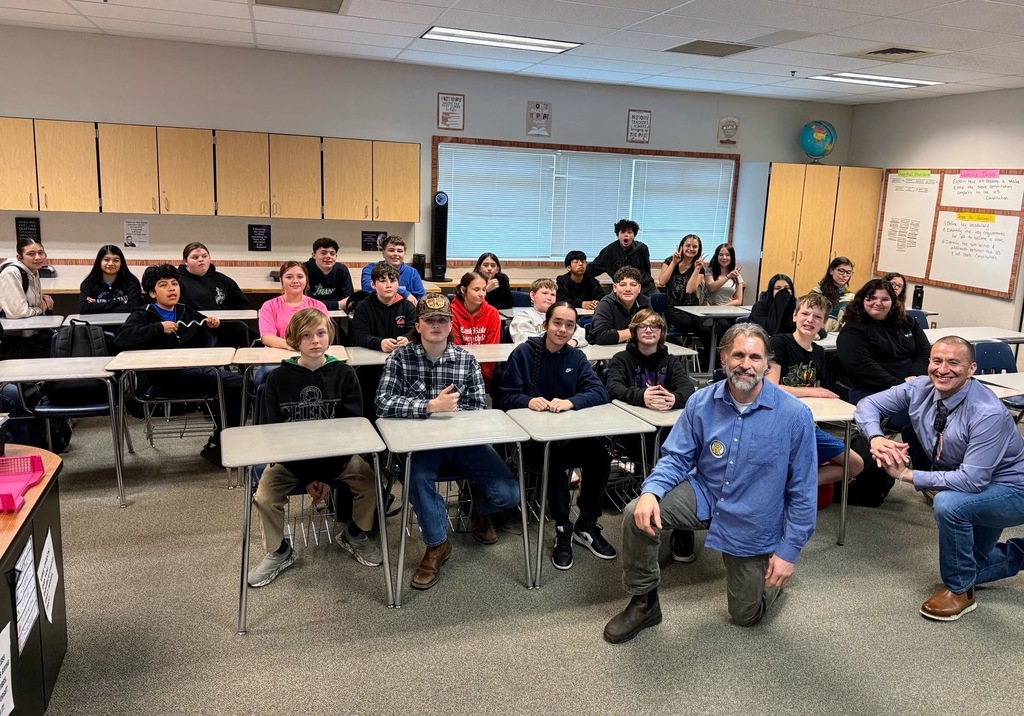 A classroom with students and teachers seated around desks. A man crouches at the front.