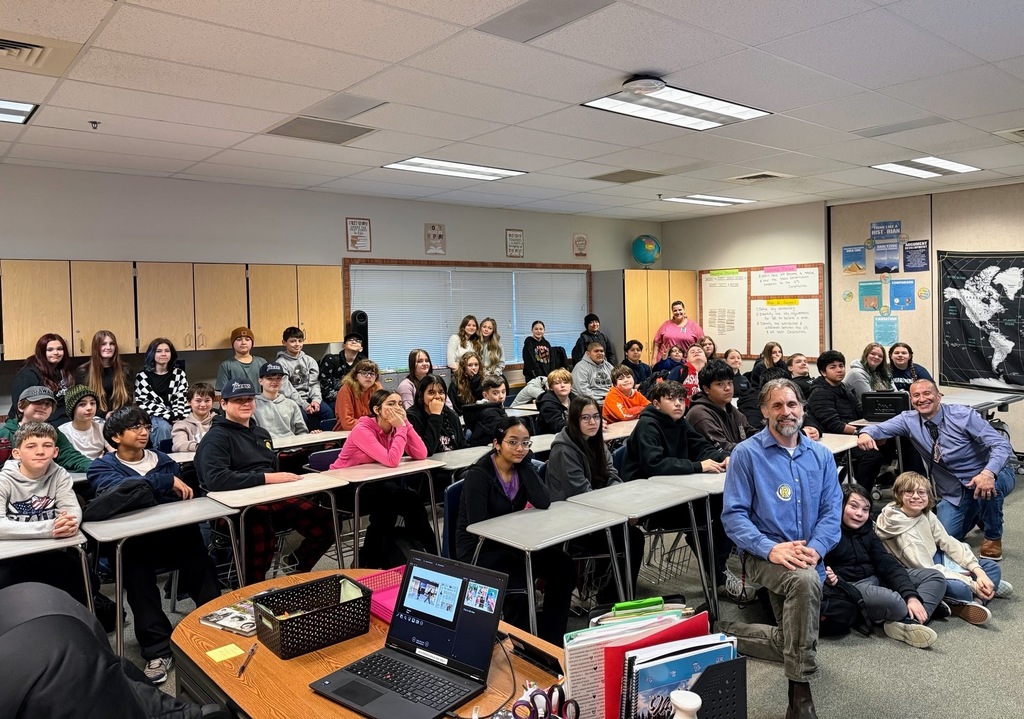 A classroom with students sitting at desks, a man standing in front, and a laptop on a table.