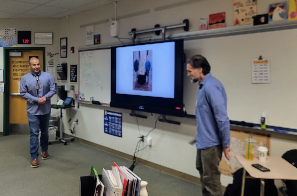 Two men standing in a classroom, one watching a large monitor displaying an image, the other near a table.