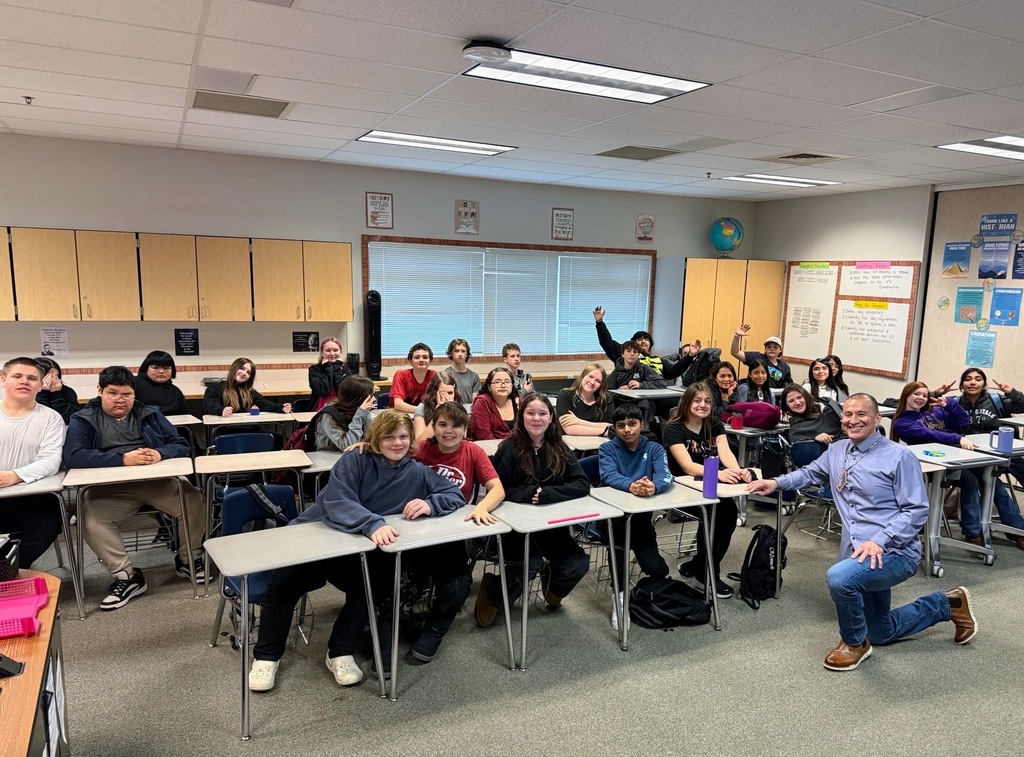 A classroom with rows of students seated at desks, one raising a hand, and a teacher kneeling in front of them.