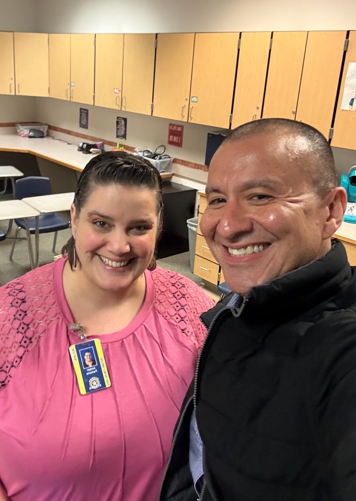 Two smiling individuals stand in a room with wooden cabinets and desks; a woman wears a pink blouse and a nametag, and a man in a black jacket.