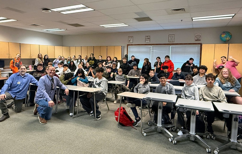 A classroom with students seated at desks, some standing, and two instructors at the front. The room has cabinets and windows.