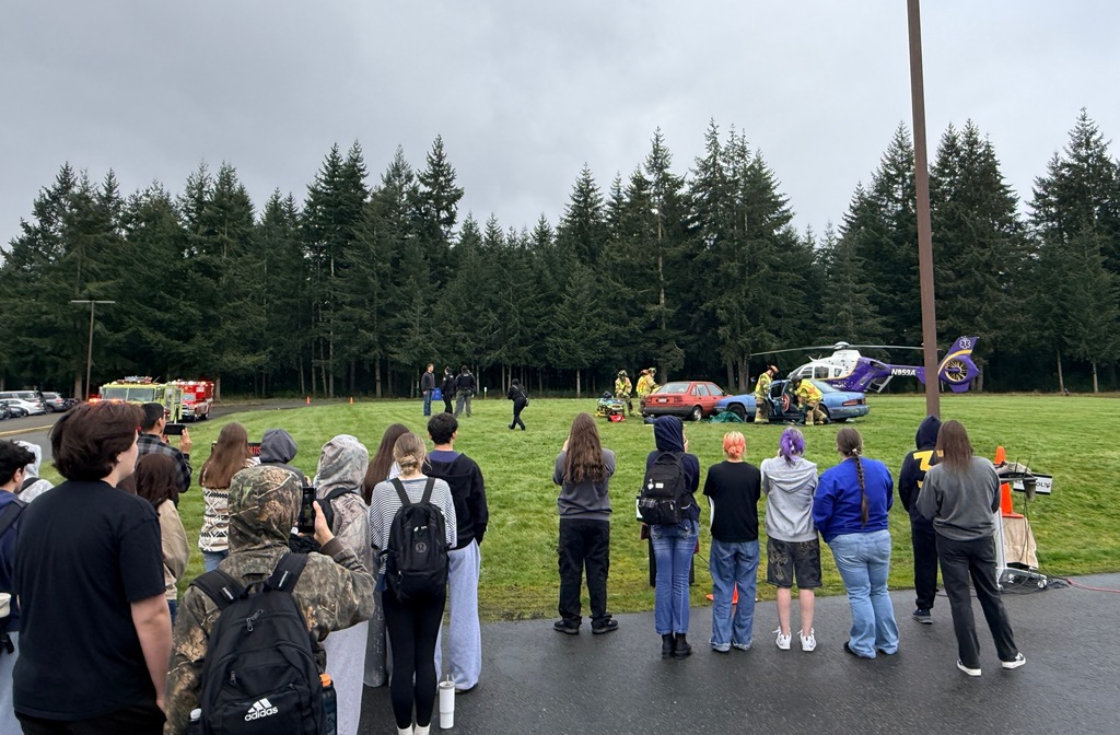 A crowd of people are standing on the road. Some wear backpacks. A helicopter is parked on a grassy field with trees in the background.