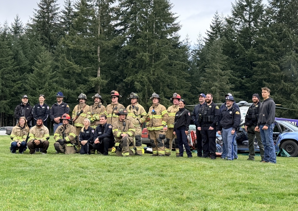 Group of firefighters in uniform posing for a photo in a grassy field with pine trees in the background.