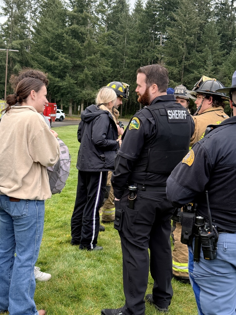 A group of uniformed people, including firefighters and police officers, are conversing on a grassy area, with trees in the background.