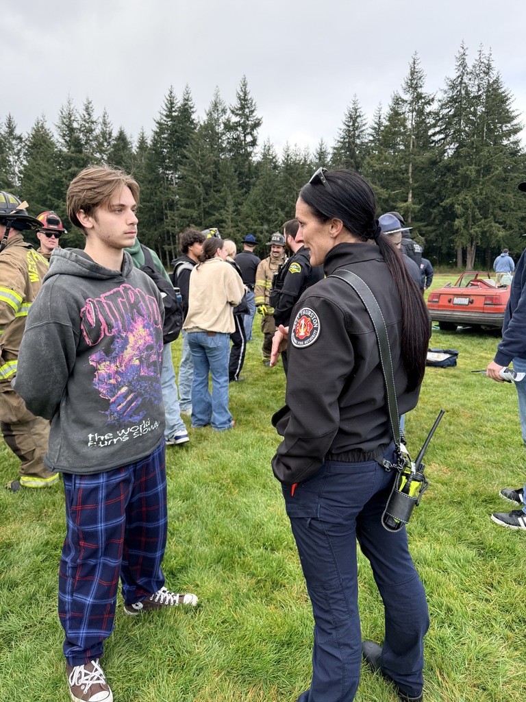 People gathered on a grassy field with trees in the background; two individuals in conversation.