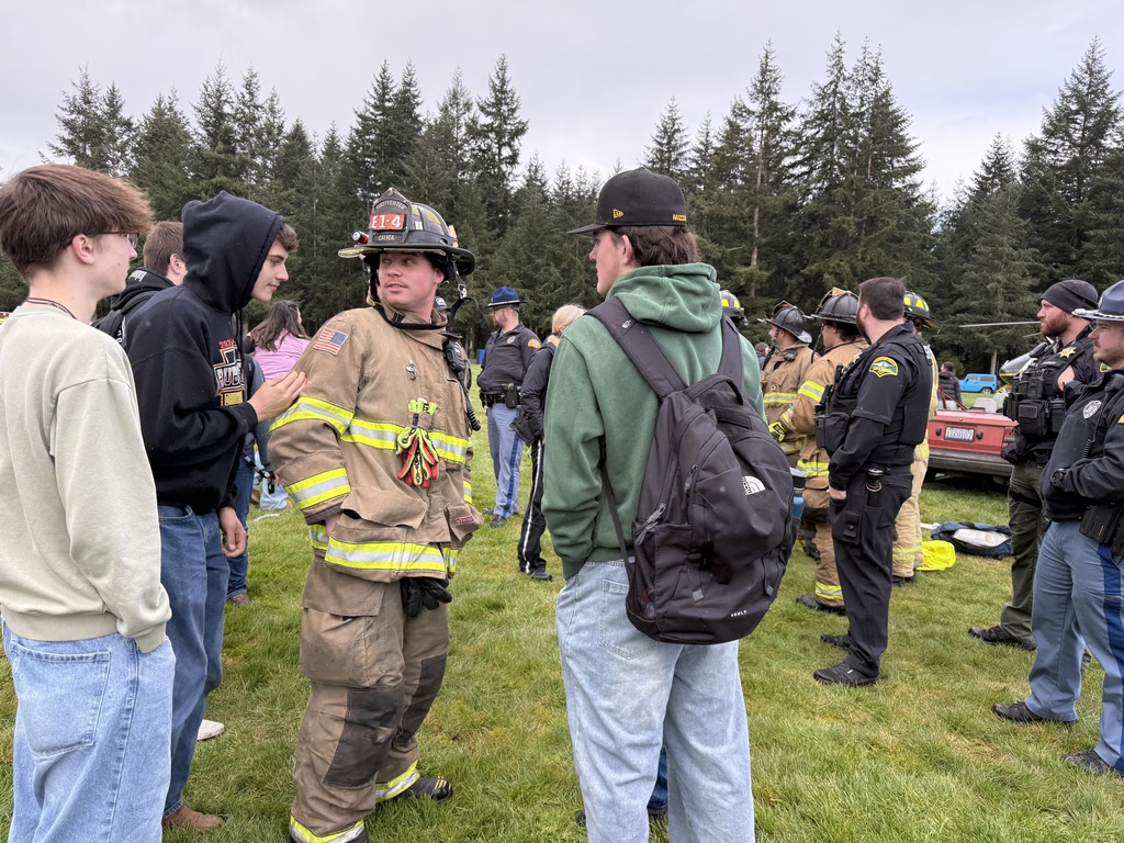 A group of people in various clothing stand in a grassy area, with trees in the background. One man wears a fireman's uniform.