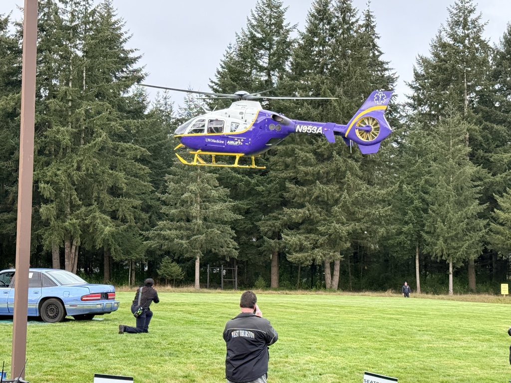 Two people on a grassy field, one kneeling. A helicopter is landing. A car is parked, and trees surround the area.