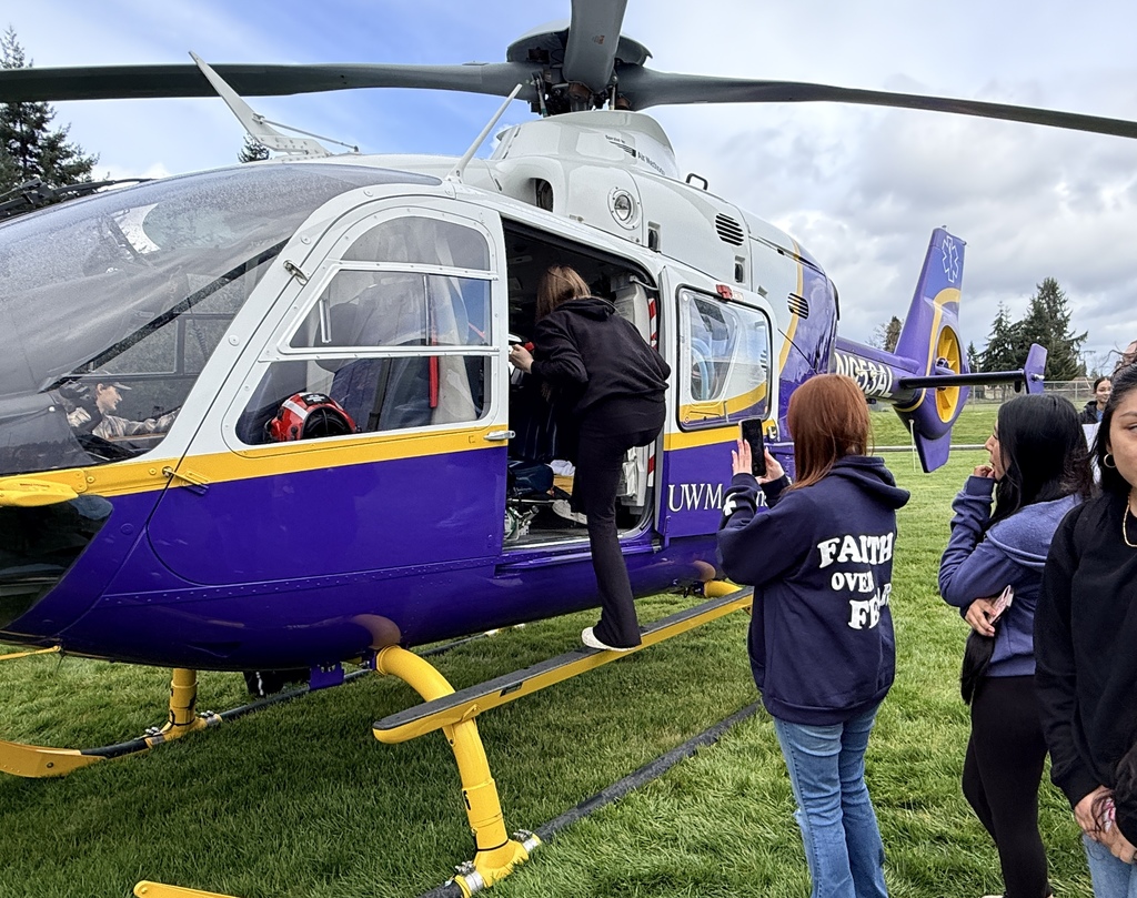 A helicopter with purple and blue colors, landing on a grass field. People watch as someone climbs inside.