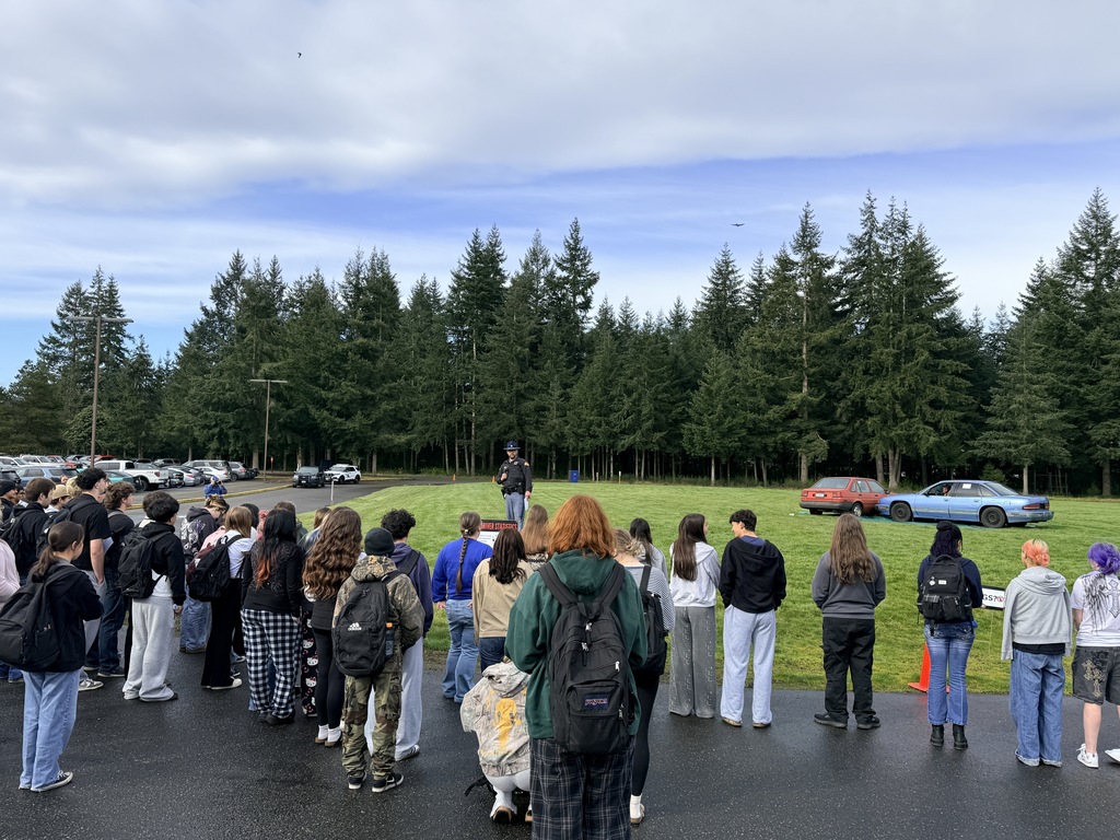 People gather on a paved area, with a person on a platform at the front. Trees and a car are in the background.