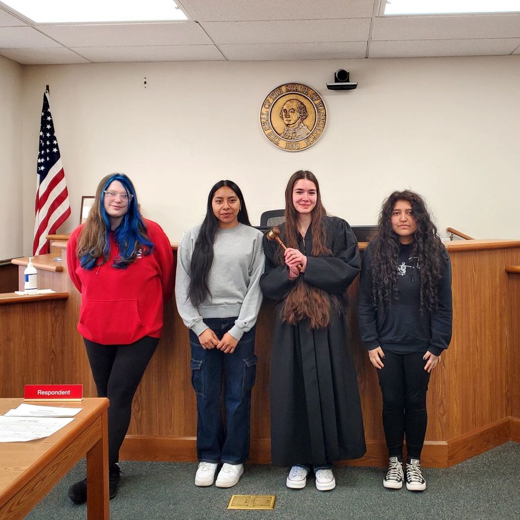 Four girls in various outfits stand in a room with a wooden counter, flag, and plaque.