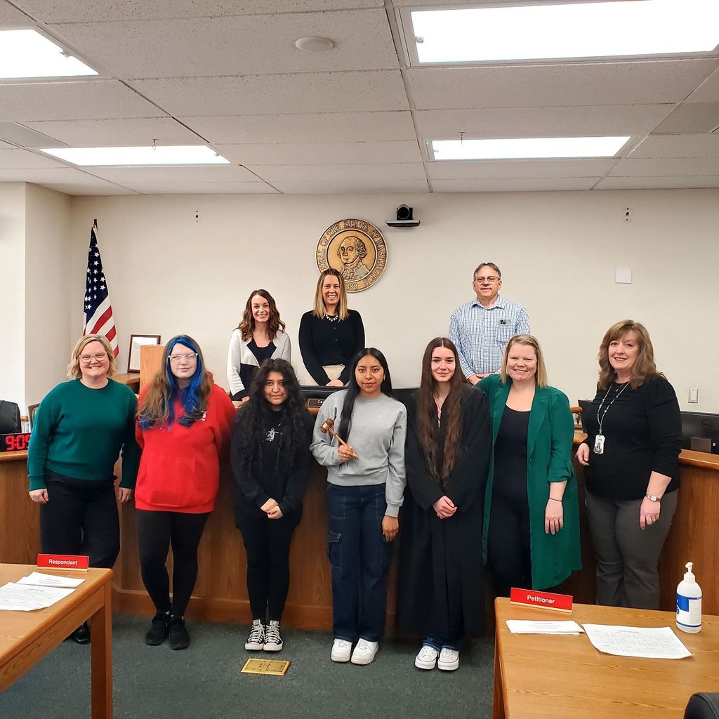 A group of nine individuals, including students, stands in a room with wooden desks and a flag.