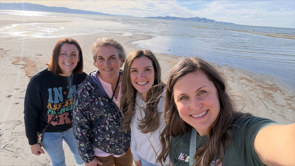 Four women stand on a beach. The woman on the right takes a selfie. Behind them is water.