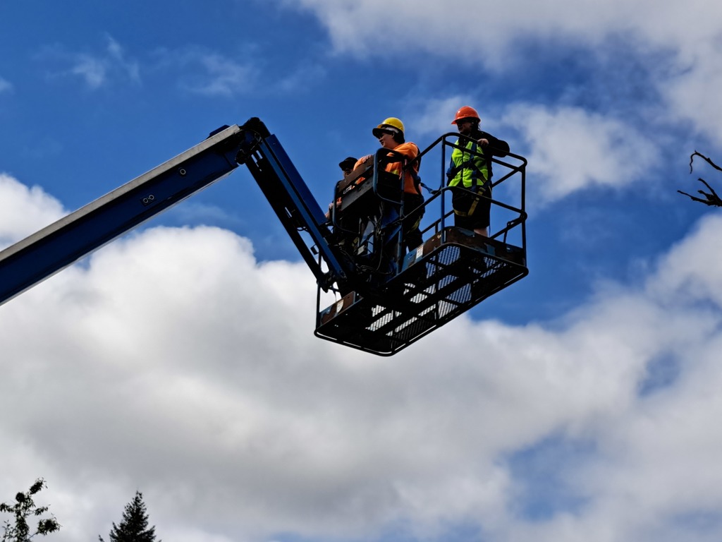 Three workers in a cherry picker on a sunny day. Two are in hard hats, one wears a reflective vest.