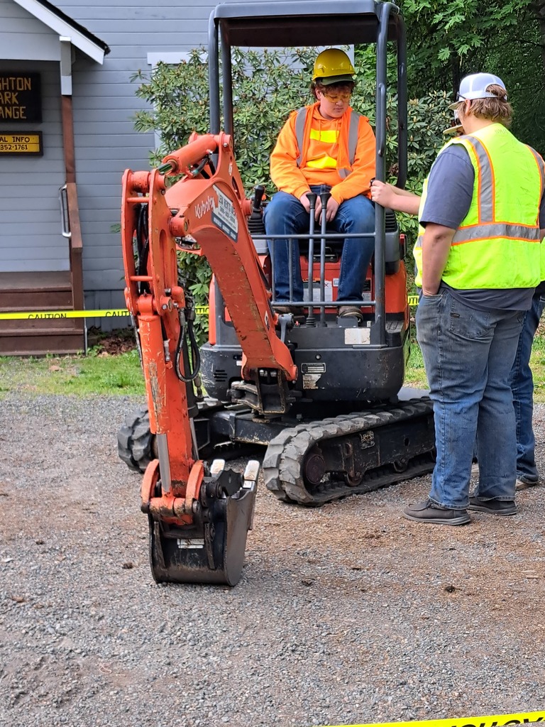 Two people in safety vests stand by a red excavator. One person is seated on it. A building is in the background.