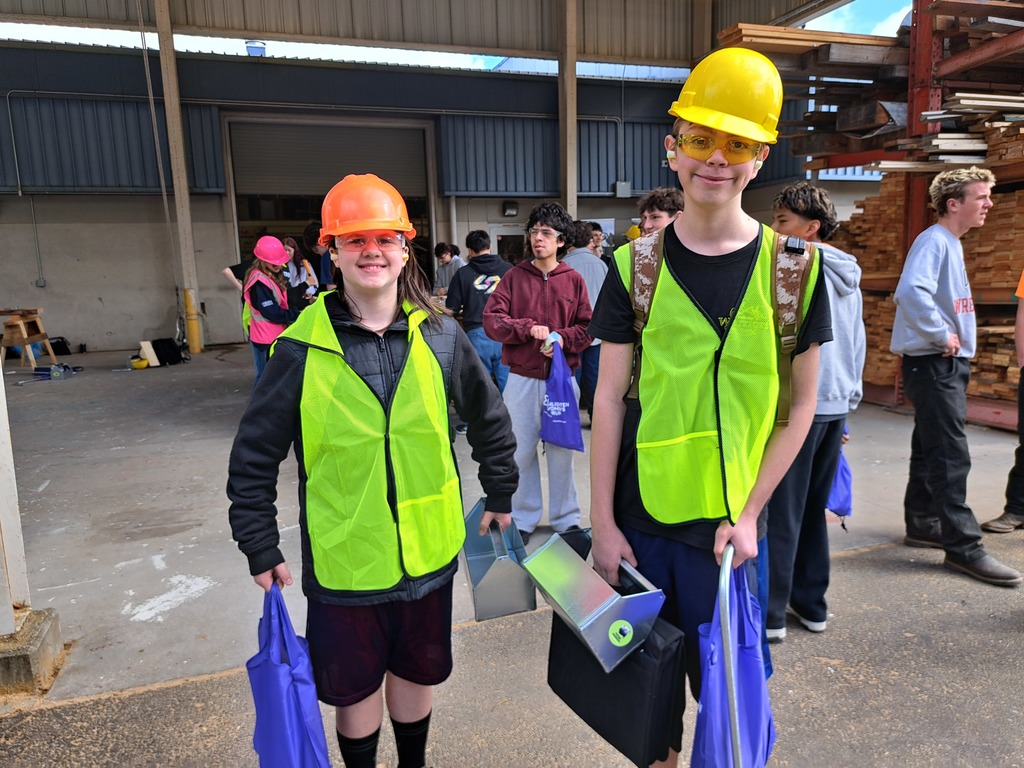 Two people in construction gear, including hard hats, stand in front of a building. They hold a tool box and bags.