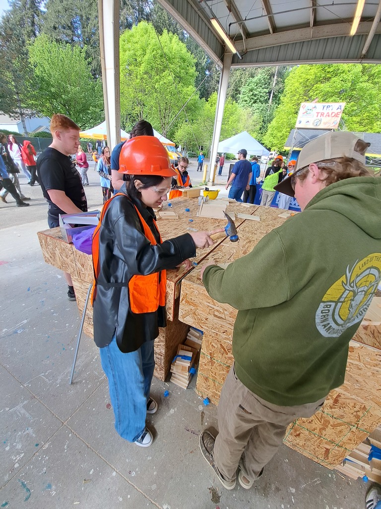 People are at a construction site, working on a project. A woman in a jacket and hard hat stands next to a man in a green hoodie.
