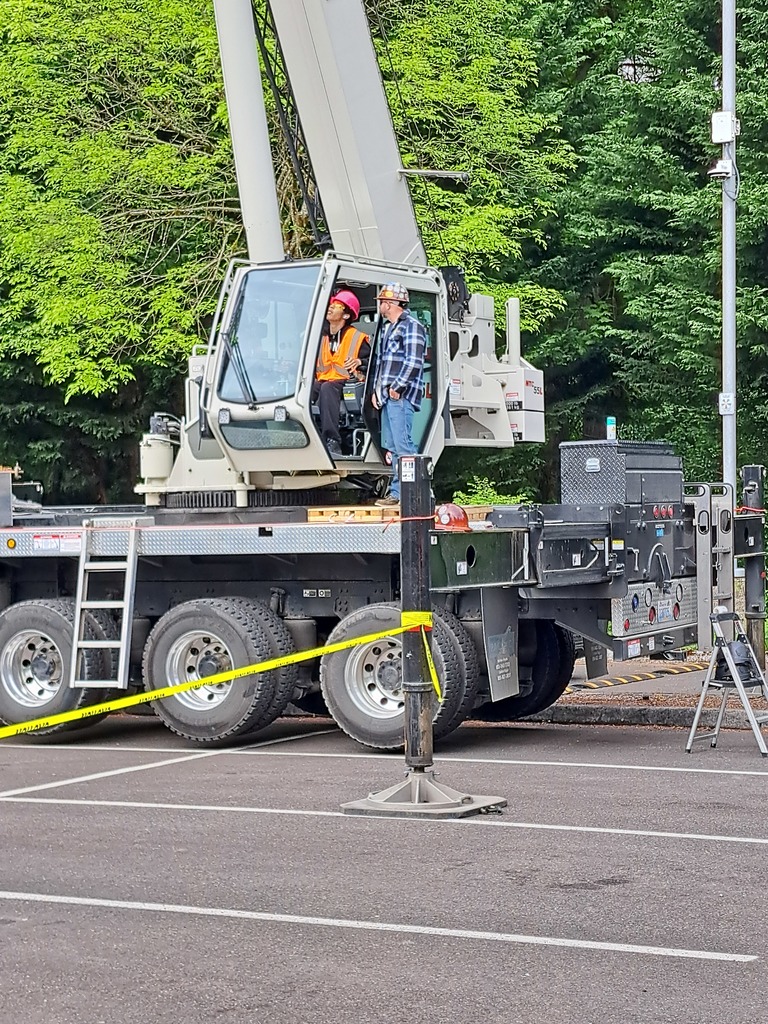 Crane vehicle parked in a paved area with two people inside. Yellow caution tape marks the ground near the vehicle.