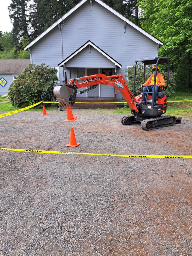 Orange excavator operating in a gravel area with orange traffic cones. Yellow caution tape is placed in the background.