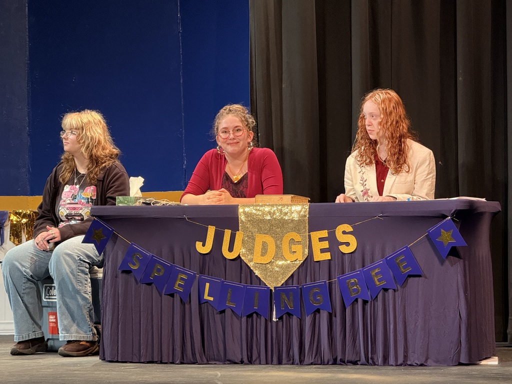 Three women sitting at a table. The table has a purple banner that reads "Judges" and "SPELLING BEE" on it.
