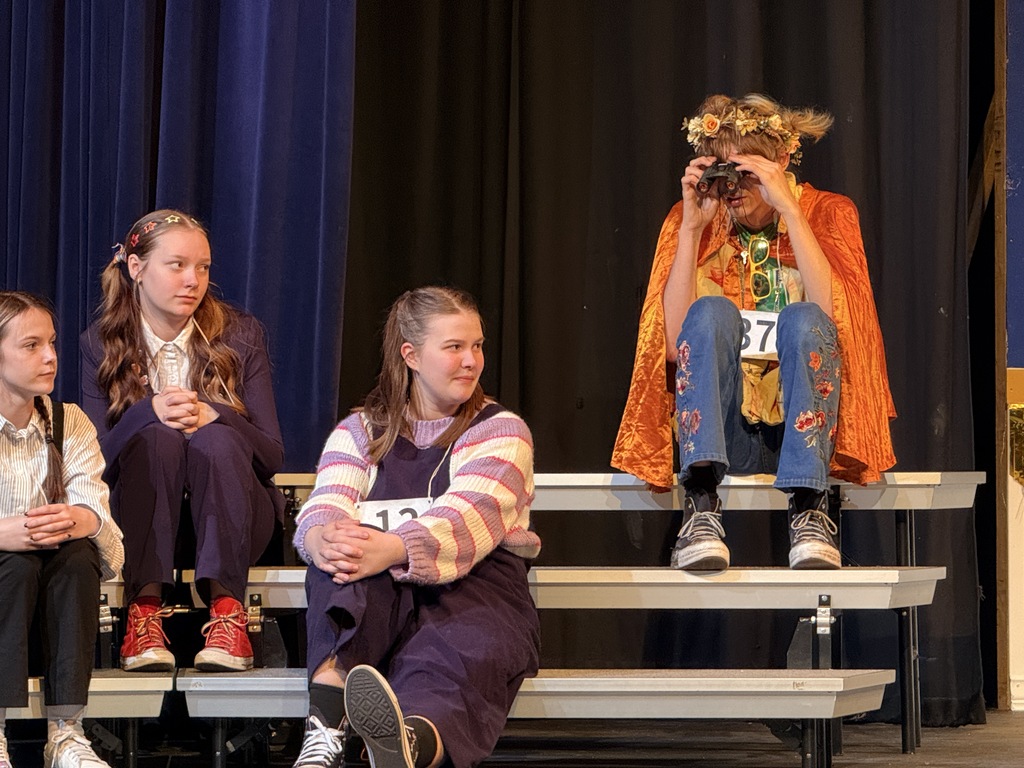 Four children, one with a crown, seated on stage with a blue curtain in the background.
