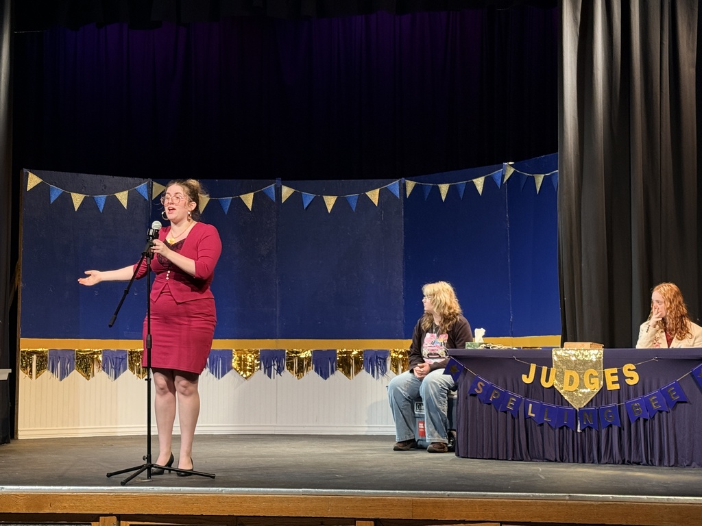 Woman in red dress speaking into a microphone on a stage; two people sit at a table behind her.