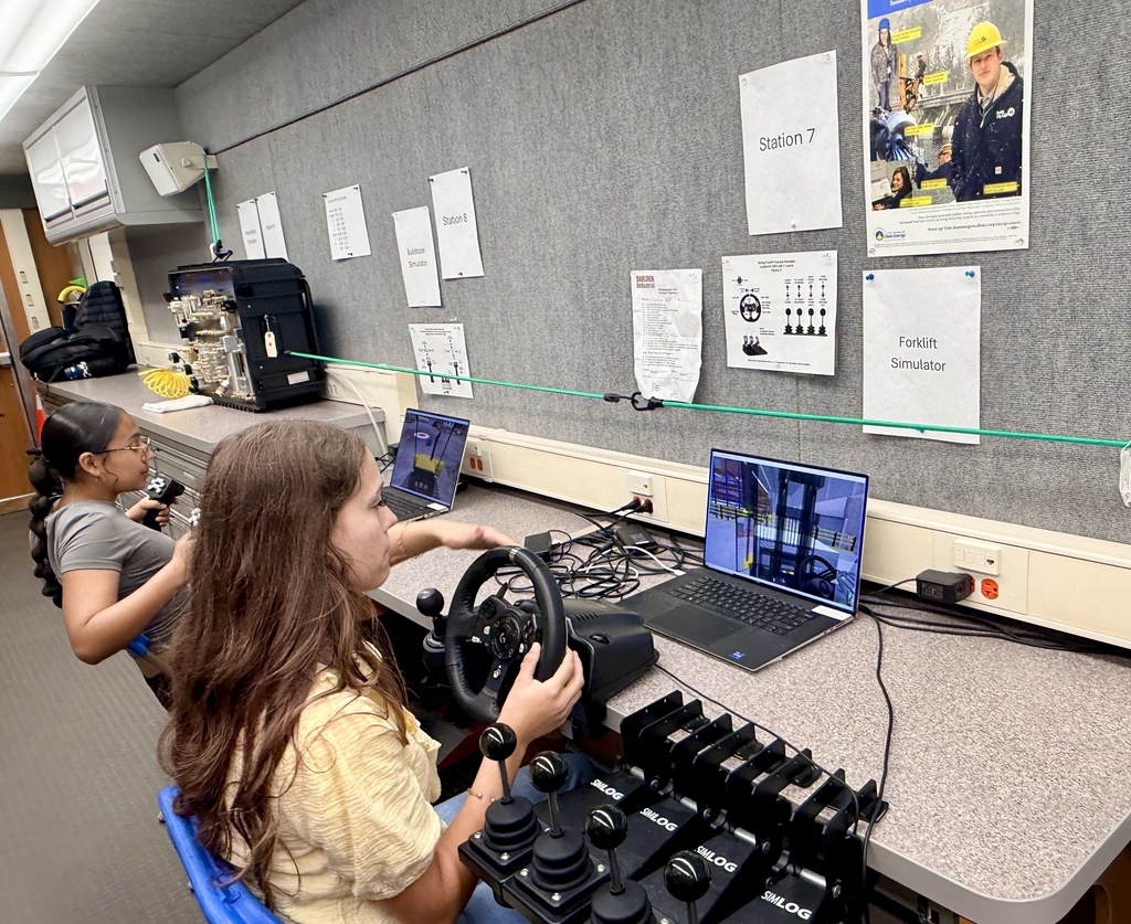 Two individuals sit in chairs, operating a steering wheel and laptop. A gray wall with papers and a photo is in the background.