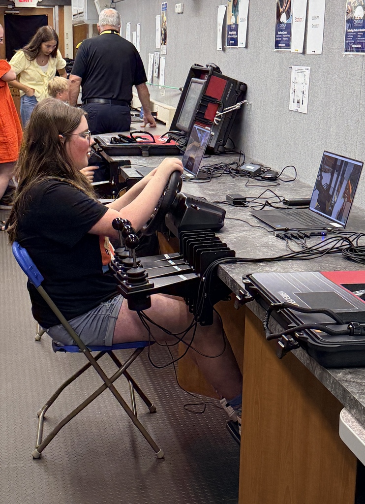 Person seated in a chair, operating a steering wheel, surrounded by monitors, laptops, and cables on a counter.