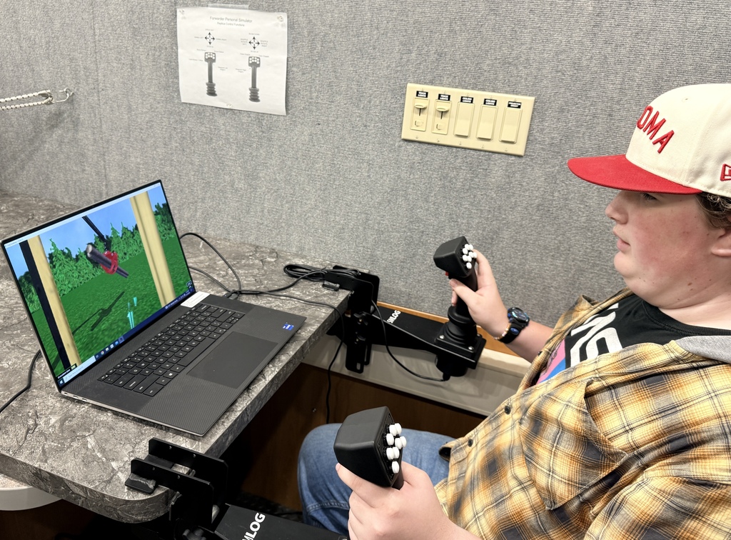 A person in a cap sits at a desk, using a laptop and a simulation controller.