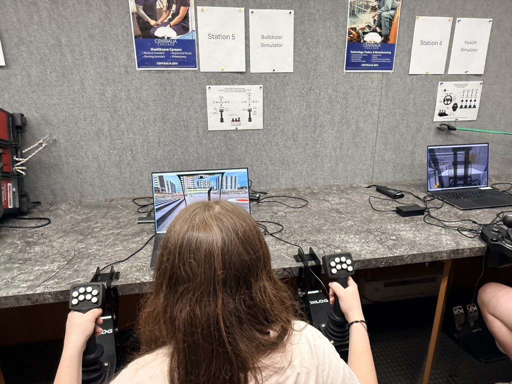 A person sits at a table, using a controller to interact with a computer display. A gray wall is behind them, with posters and various items attached.