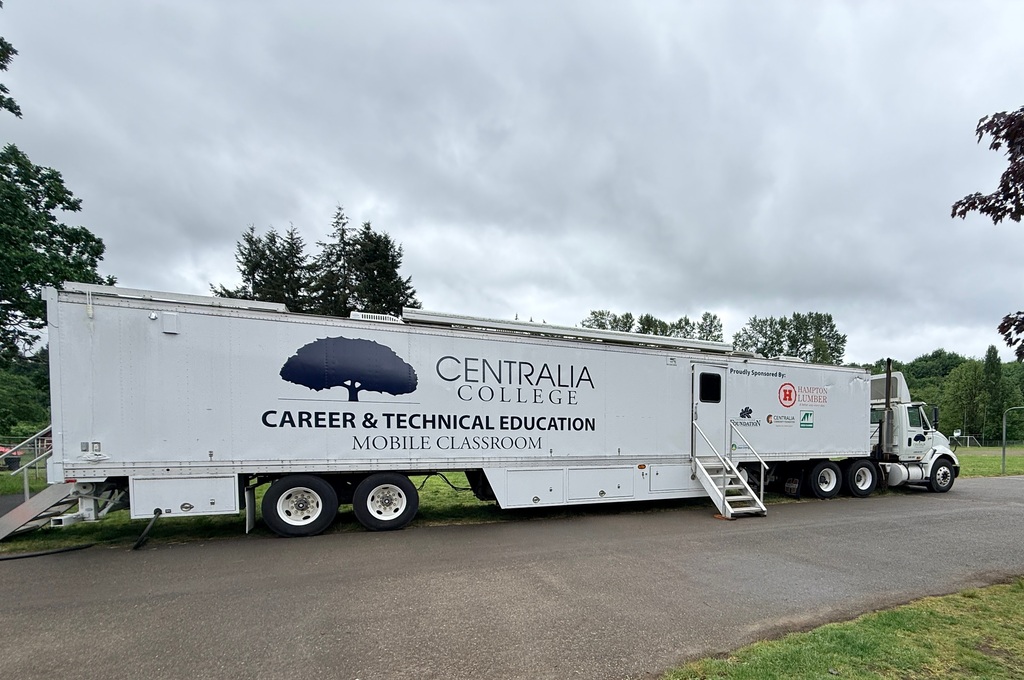 White trailer with "Centralia College Career & Technical Education Mobile Classroom" text parked on road with trees and cloudy sky.