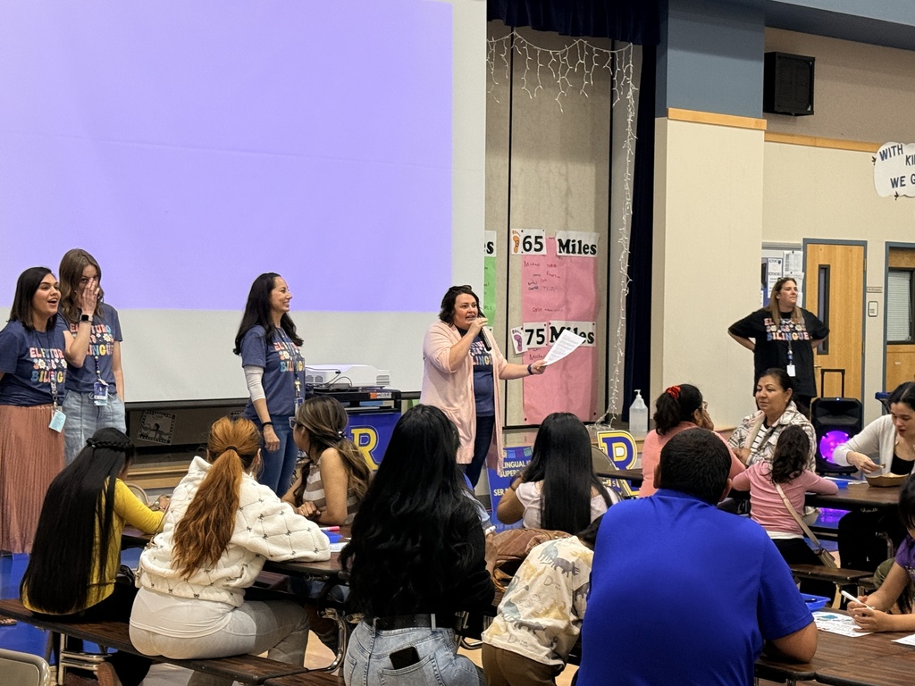 Several people stand in front of a projector screen while others sit in chairs. A woman speaks to the group.