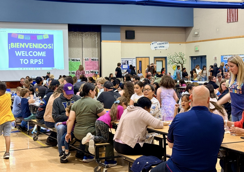 Large group of people seated at tables in a school cafeteria. Overhead projector displays "Bienvenidos! Welcome to RPS".
