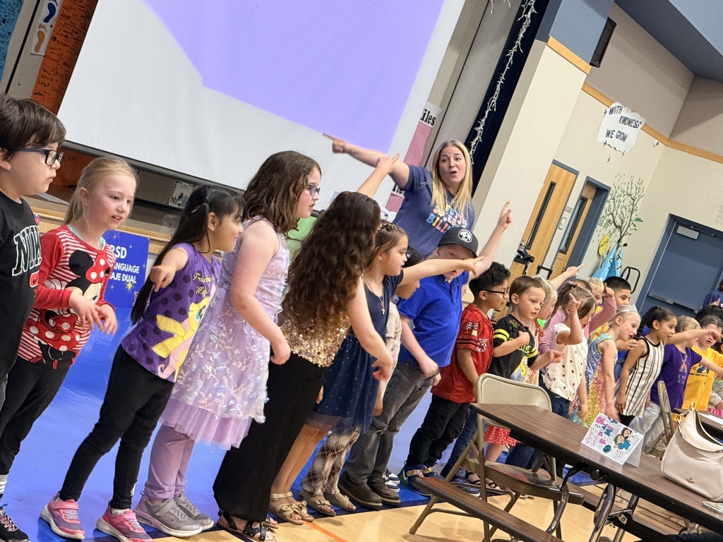 Group of children standing in a school gym with an adult; one child holds a microphone.