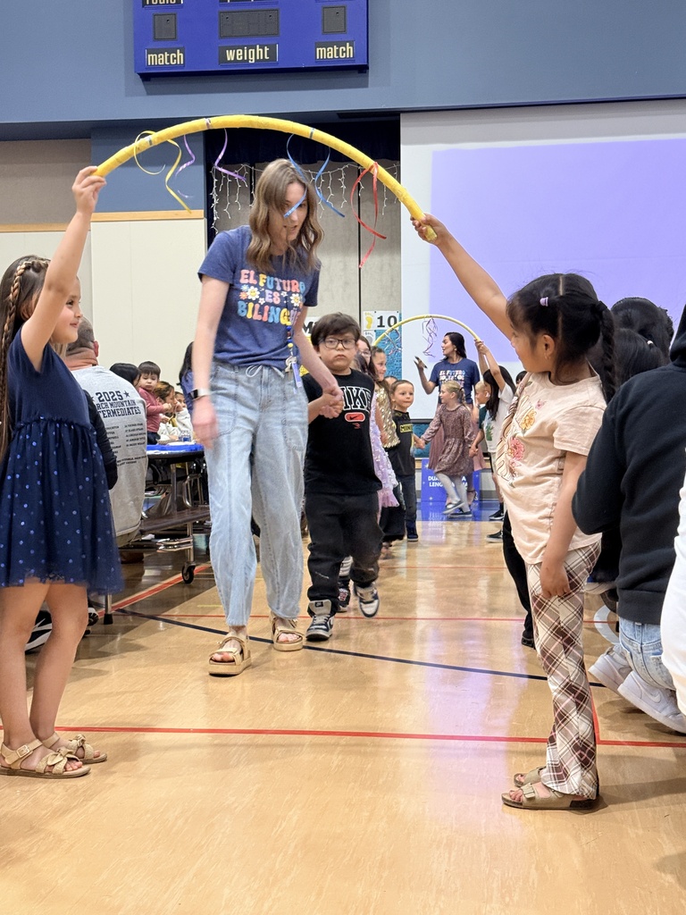 People gather in a gymnasium, a woman holding a yellow rope, while children and adults walk around, with a scoreboard in the background.