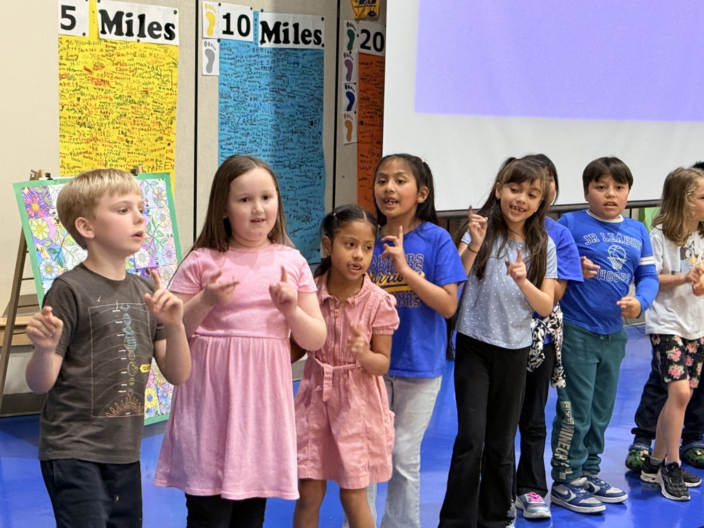A classroom with children in various outfits standing in a line, one gesturing. In the background, colorful posters with "5 Miles" and "10 Miles" are visible.