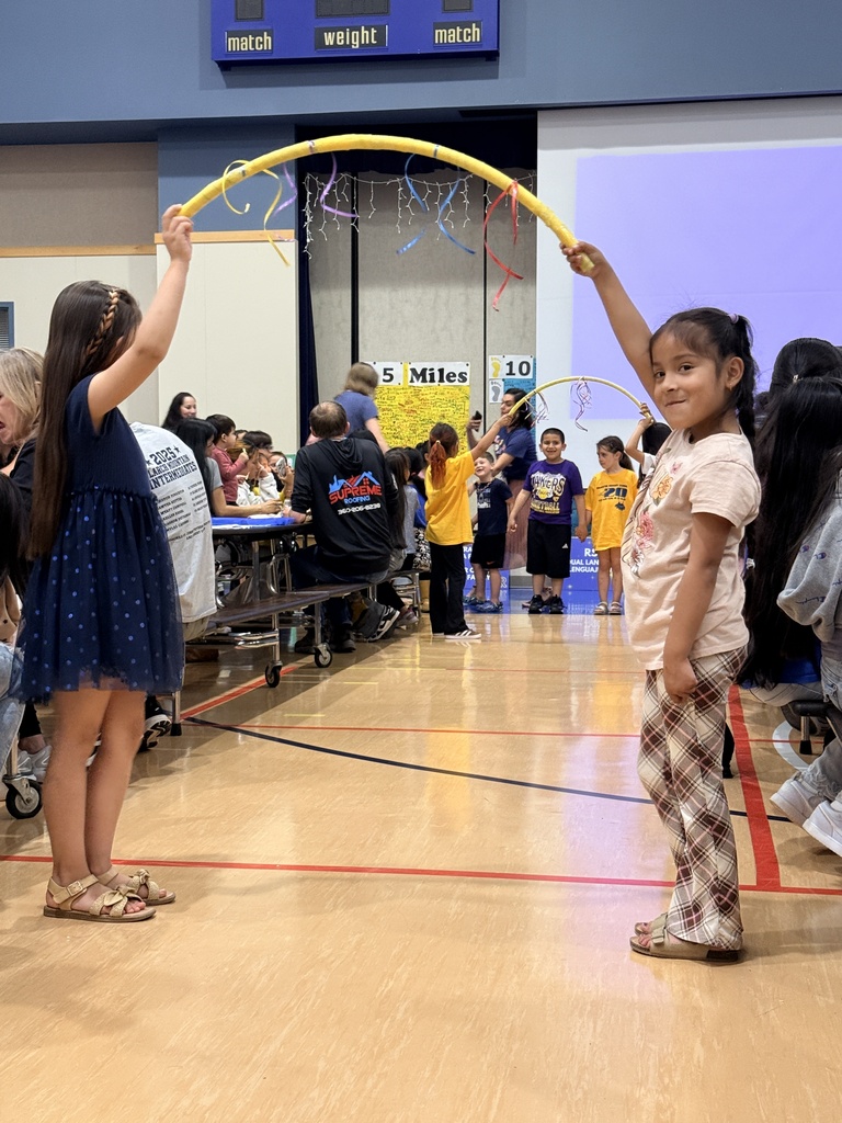Two girls play with a long, yellow hula hoop in a gymnasium with others seated and standing.