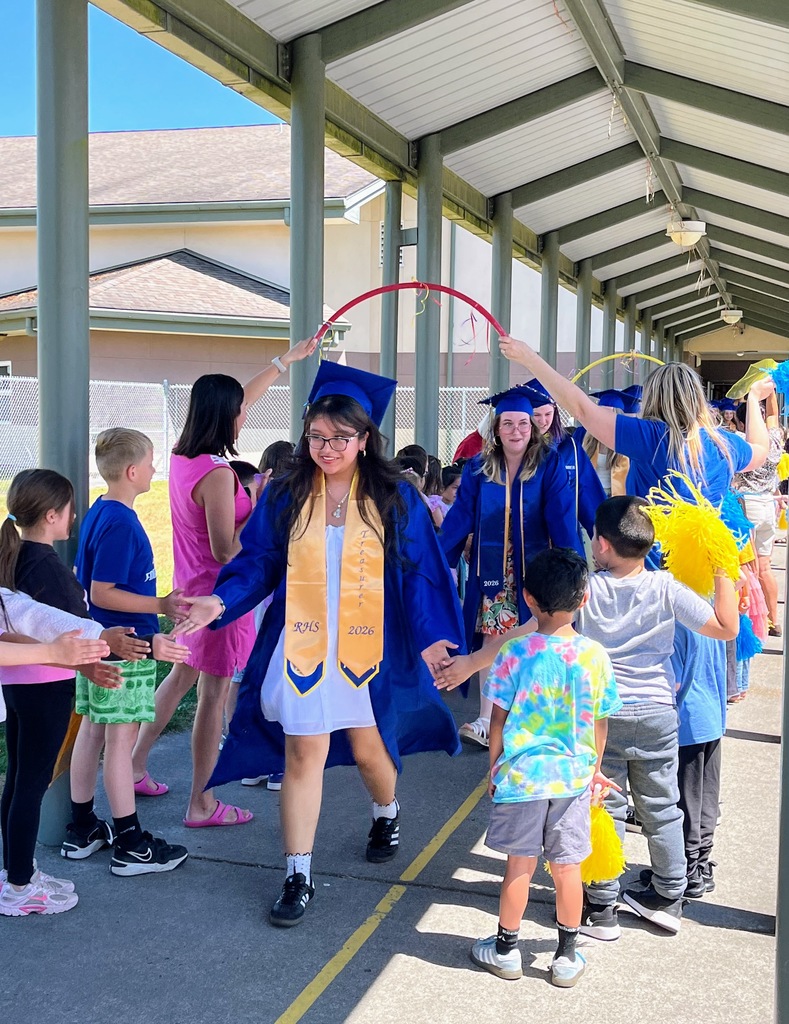 A group of graduates walk through a school corridor, holding hands. Some wear caps and gowns, and one holds a hula hoop.